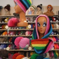 Schaufensterpuppen tragen selbstgestrickte, bunte Schals und Mützen vor einem Verkaufsregal, gefüllt mit Wolle